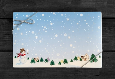 Blue snowman box covered with white snowflakes on a blue and white background with a two story tan house in the background on the right of the box. Young evergreen pine trees adorn the foreground.. Photo taken on black wooden background.